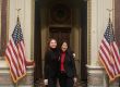 Cathy Cheng and Gabrielle Beck standing together, arms around each other, at the entrance to the Executive Office Building. Fancy tiles floor, arched doorway, America flags flanking.