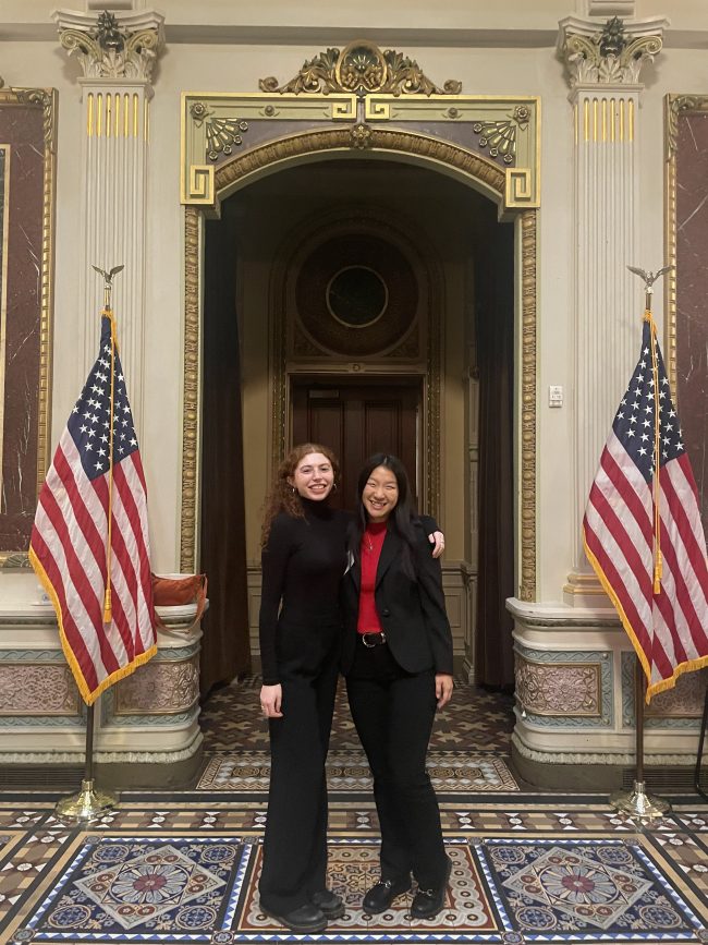 Cathy Cheng and Gabrielle Beck standing together, arms around each other, at the entrance to the Executive Office Building. Fancy tiles floor, arched doorway, America flags flanking.